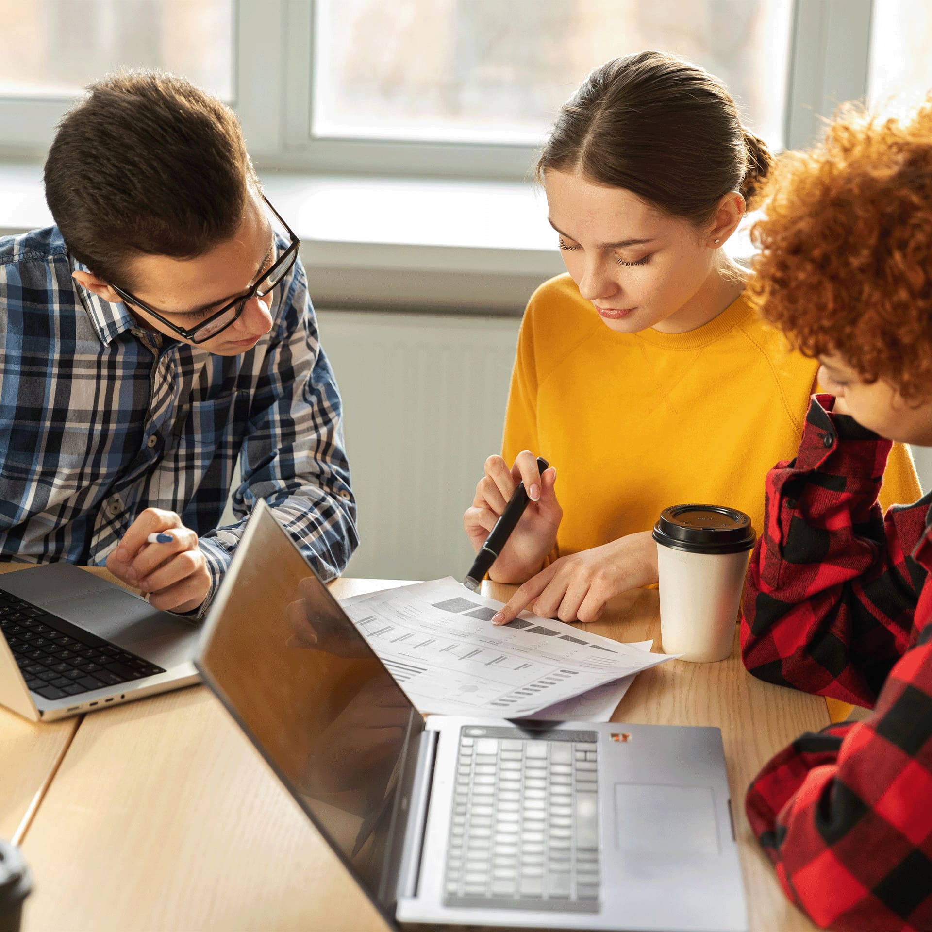 Three young professionals working on their finance apprenticeships in a well-lit office space, actively engaged in discussion over documents and laptops.