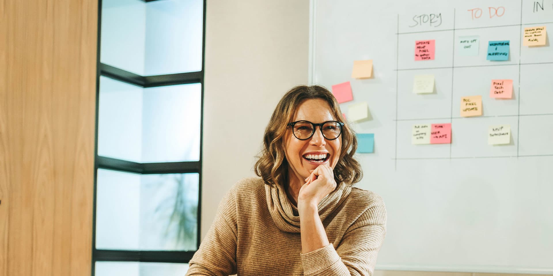 A woman wearing glasses is sitting at a table with a mug, holding a pen and smiling.