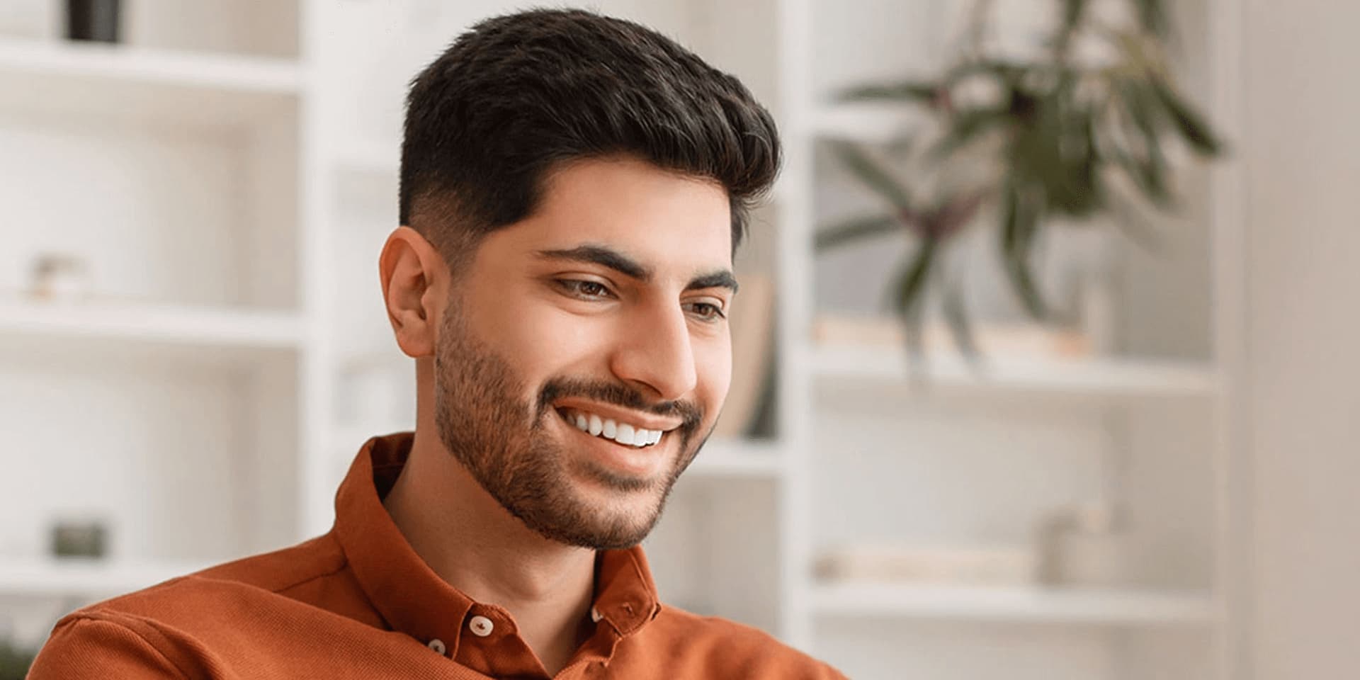Young smiling man wearing an orange shirt, typing on his laptop.