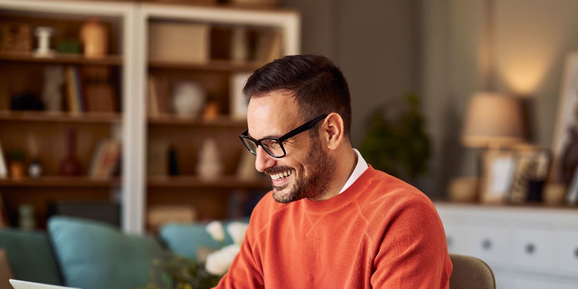 A man wearing an orange sweater and glasses is sitting at his desk, working and smiling.