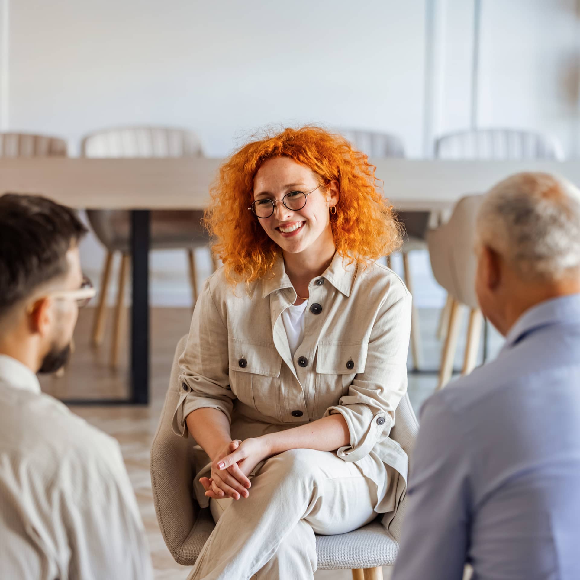 Woman with curly red hair in cream shirt speaks with two men about mental wellbeing in a modern office.