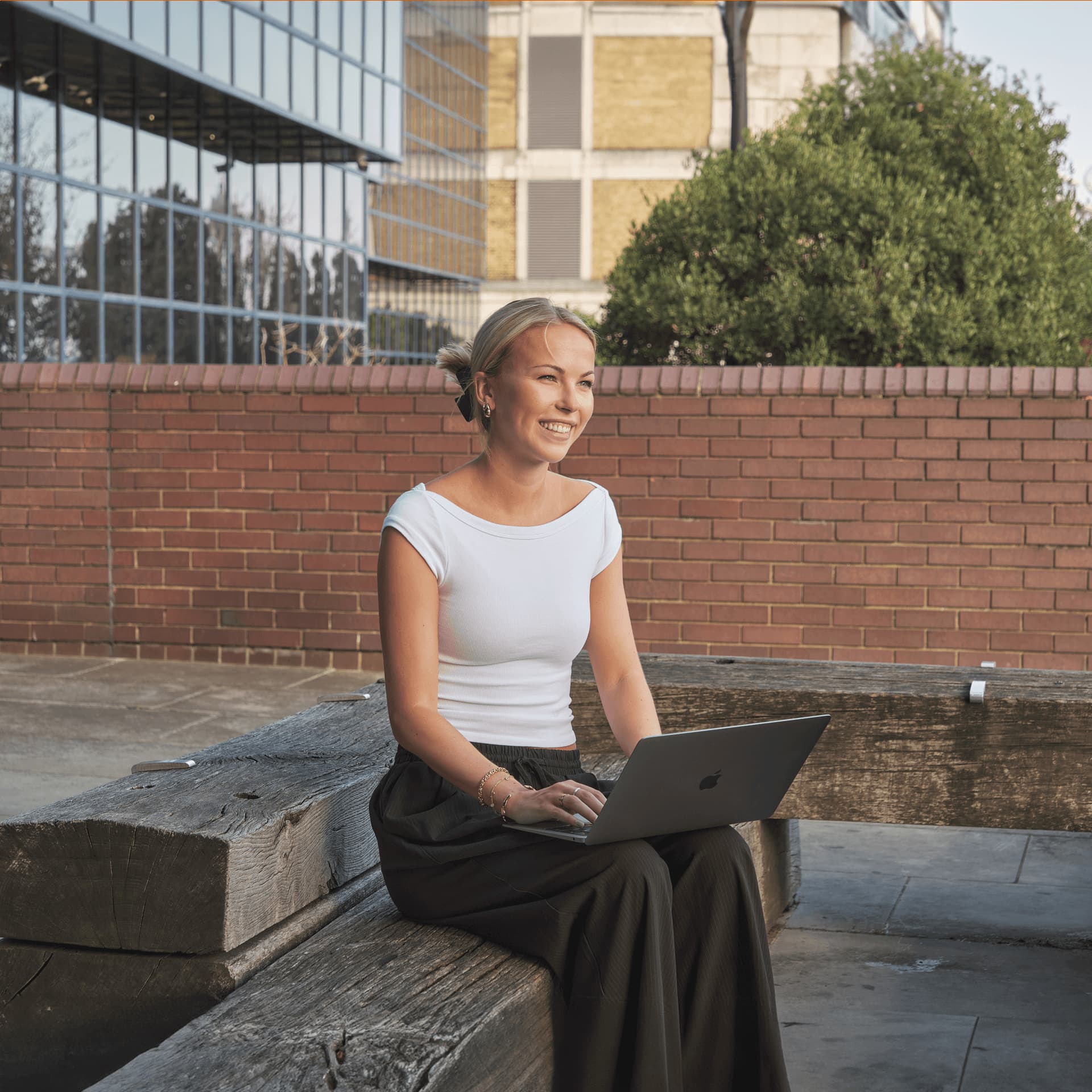 Smiling female student sits outside on laptop preparing for online assessment.
