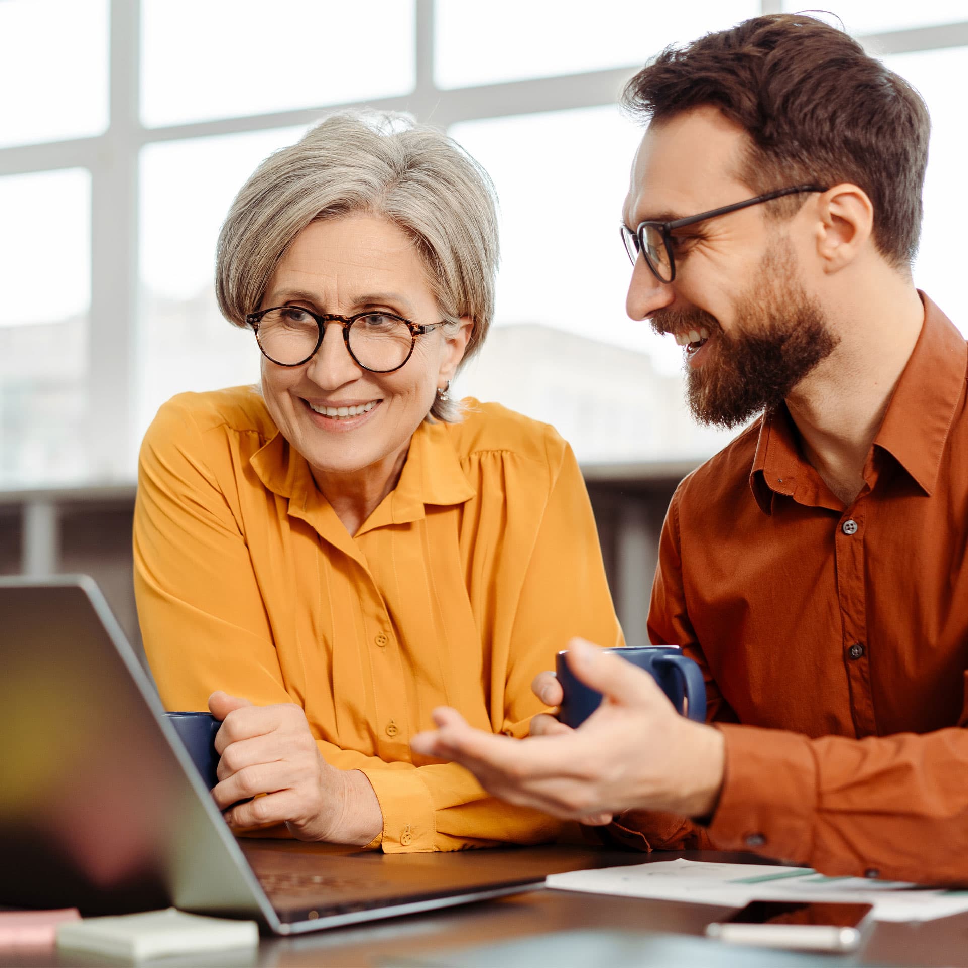 A man and a woman holding mugs, looking at the laptop and smiling.