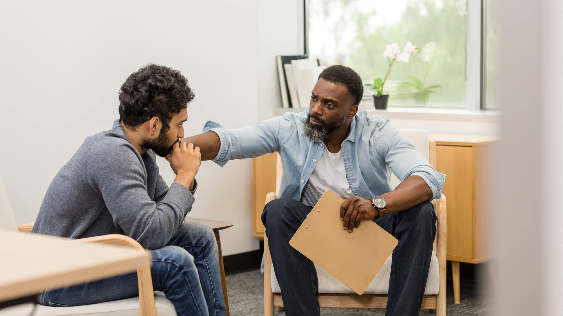 Male therapist in blue shirt has hand on shoulder of man seeking therapy.