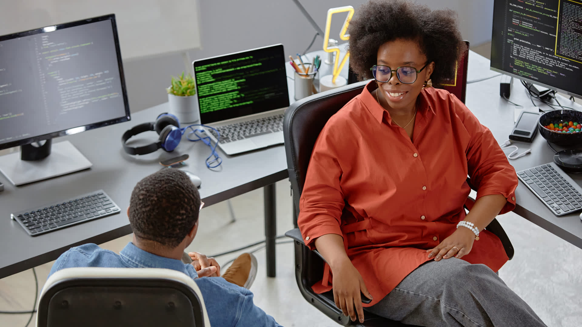 Female cloud architect in orange shirt works on programming at a desk with a colleague.