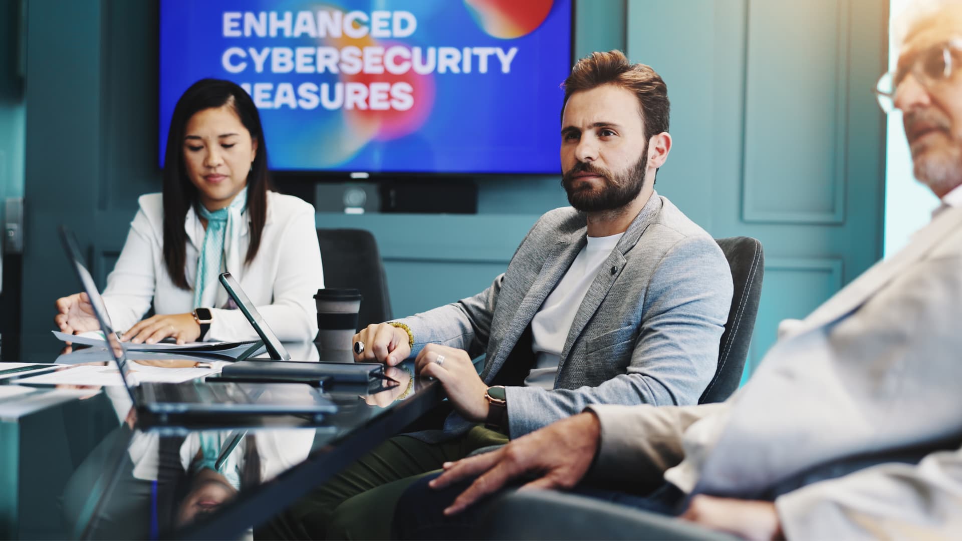 Man in grey blazer sat with colleagues in a meeting about cyber security.