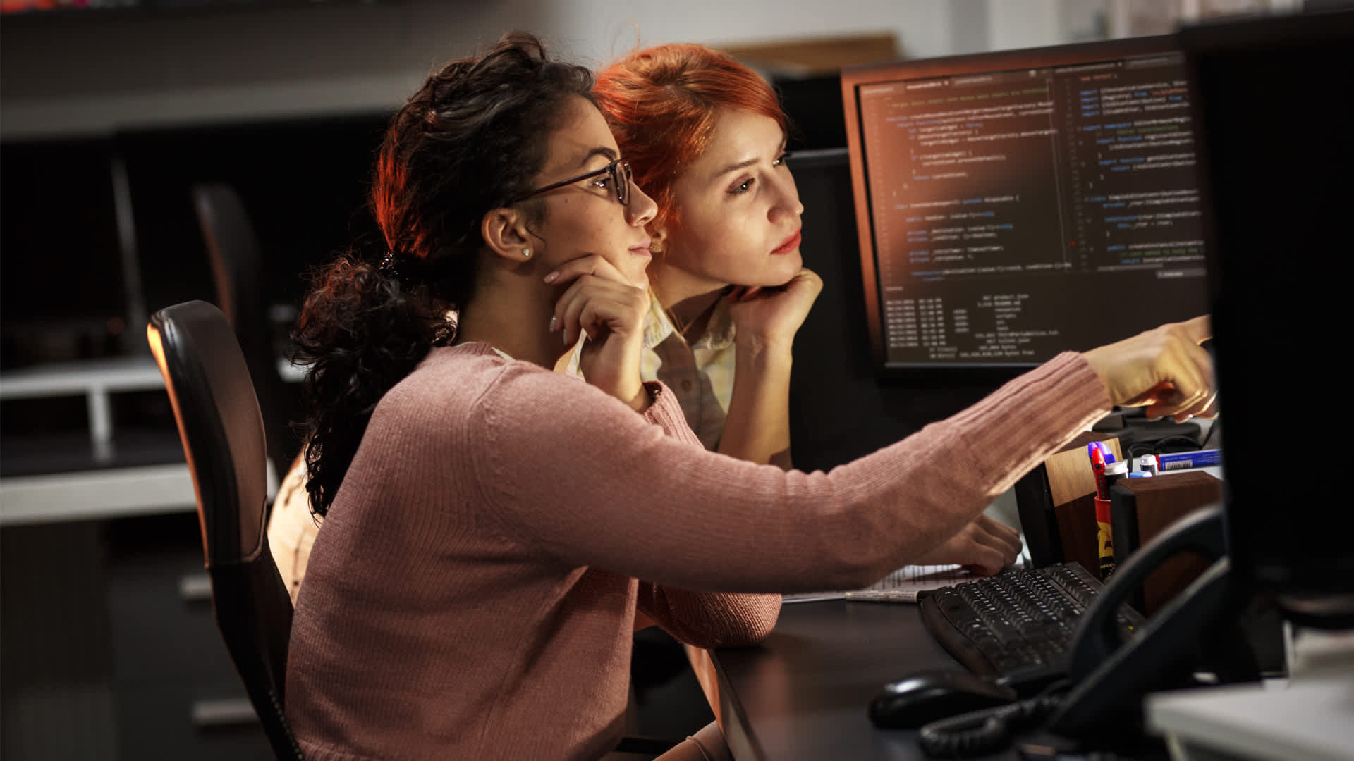 Two female colleagues use object oriented programming in penetration testing role in dark office.